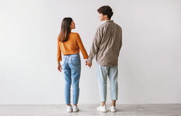 Full length of young Asian man and woman standing with their backs to camera, looking at each other and holding hands against white studio wall. Millennial couple expressing affection