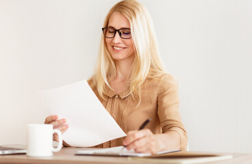 Poster - A woman with blonde hair and glasses sits at a desk, reviewing documents and taking notes.