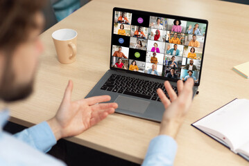 Poster - A man sits at a desk in front of a laptop, actively participating in a video conference meeting with colleagues. His hands are raised, gesturing and engaging with the online participants.