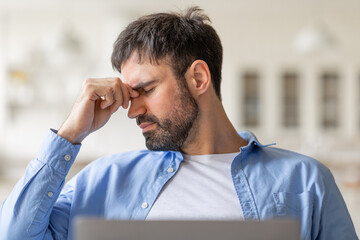 Poster - Tired man sitting in a chair inside, looking down and rubbing his eyes with his right hand. He appears tired or stressed, and is likely working on a laptop computer.