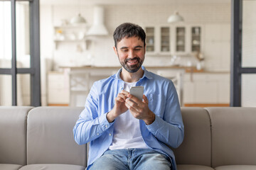 Poster - A man wearing a light blue shirt and jeans sits on a couch in a living room, smiling as he uses a smartphone. There are white cabinets in the background and a large window