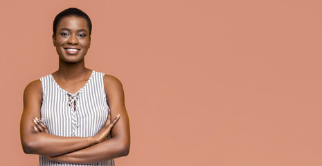 Wall Mural - This image features a smiling African American young woman with short black hair, wearing a striped sleeveless dress, standing against a peach-colored background with her arms crossed.