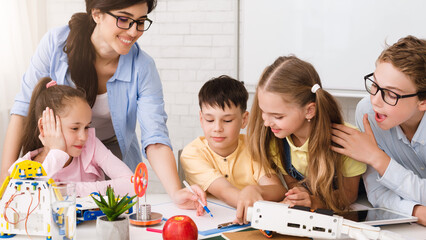 Poster - A group of elementary school students are engaged in a STEM learning activity in a classroom setting. They are working with robots and building models, and a teacher is guiding their learning