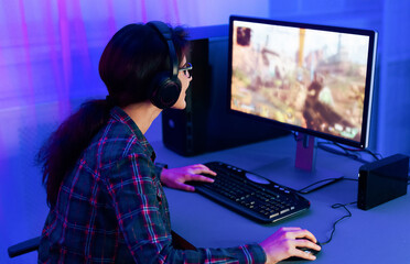 Aufkleber - A young woman sits in a chair in front of a computer, wearing headphones and playing a video game in a room lit in blue light. Her hands are on the keyboard and mouse.