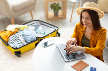 Happy young woman planning vacation travel with laptop pc, reading tourist blog online, booking tickets or hotel room on web, getting ready for abroad journey at home, free space