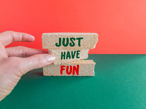 Concept words 'Just Have Fun' on the brick blocks. Beautiful red background, green table. Businessman hand. Inspiration quote.