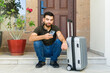 © AntonioDiaz - Young Hispanic man waiting for a taxi with his suitcase while using a ride sharing app on his phone on the front steps of his home
