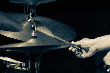 Part of a drum set with emphasis on the cymbals and the drummer's hand holding a wire brush drumstick. The photo is in black and white with selective focus.