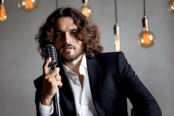 Canvas Print - Portrait of handsome attractive singer in classic suit on grey background. Man singing pop hit with open mouth in retro microphone