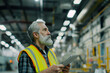 © mankjon - A mature bearded man with gray hair wearing a yellow high-visibility vest, standing in an industrial warehouse, intently looking at a digital tablet