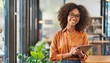 © gankevstock - A woman with curly hair and glasses smiles while holding a tablet, wearing an orange shirt in a bright, modern office indoor setting