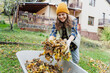 © olezzo - Young happy woman collecting autumn leaves in the backyard of a country house using a rake and cart