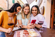 © Xavier Lorenzo - Three young women using mobile phone while enjoying appetizer in a bar. Millennial diverse italian girls chatting on smart phone while drinking red wine