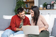 © Srdjan - Simple living Young engaged couple sitting at home, spending quality time, woman searching for wedding dress on laptop, man reading a book, organizing wedding ceremony. Searching honeymoon destination