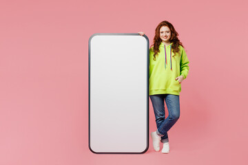 Wall Mural - Full body young ginger woman wear green hoody casual clothes stand near big huge blank screen mobile cell phone smartphone with area isolated on plain pastel light pink background. Lifestyle concept.