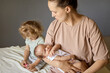 © sementsova321 - Older Caucasian sister sitting on bed in room with little sibling and mommy beautiful mother holding baby in hands eldest daughter jealous of her mother's newborn baby