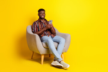 Wall Mural - Full size photo of nice young man sit armchair smart phone wear shirt isolated on yellow color background