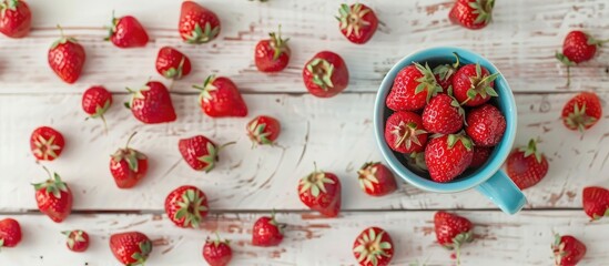 Fototapete - A blue cup holds ripe red strawberries on a white wooden table surrounded by sweet scattered berries in a top view copy space image