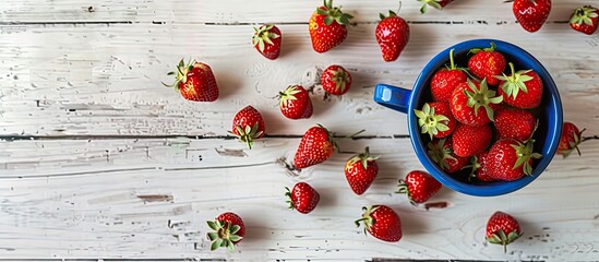 Fototapete - A blue cup holds ripe red strawberries on a white wooden table surrounded by sweet scattered berries in a top view copy space image