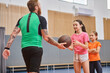 © LIGHTFIELD STUDIOS - A male teacher stands holding a basketball in front of a diverse group of children in a bright, lively classroom setting.