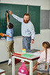 © LIGHTFIELD STUDIOS - A male teacher stands confidently in front of a blackboard, passionately educating a group of children in a bright, lively classroom setting.