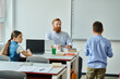 © LIGHTFIELD STUDIOS - A man sits at a desk in front of a group of children, engaging them in a lively classroom setting.