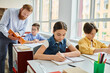 © LIGHTFIELD STUDIOS - A group of children engage in learning activities, seated at bright desks in a lively classroom led by a male teacher.