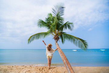 Alone woman traveler standing under a palm tree while relaxing on a tropical sandy island and enjoying idyllic and beautiful moment life