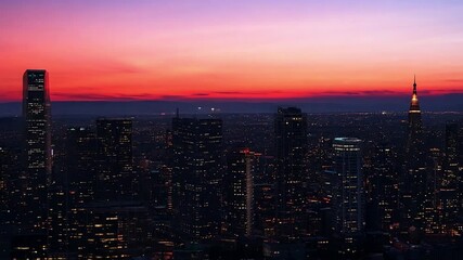 Poster - A panoramic view of a city skyline at sunset with a colorful sky. Tall buildings with lights on are seen in the foreground, while the city extends into the distance.