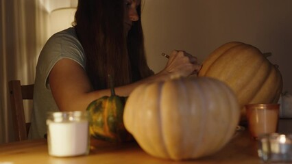 Wall Mural - A Long-Haired Brunette at a Wooden Table with Various Pumpkins Draws Contours for Carving Jack-O'-Lanterns for a Halloween Party.