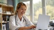 © ifoto - Czech female doctor working on laptop in a clinic, smiling and looking at the screen while sitting behind a desk with a stethoscope