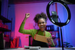 © amnaj - Young woman happily records content for her online business in her vibrant home office, holding a package and surrounded by greenery