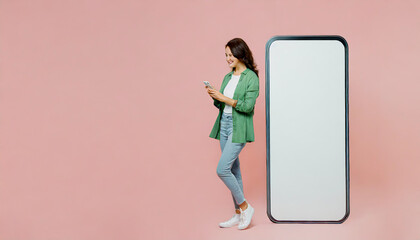 Full body happy sideways young woman in knitted sweater sit near big huge blank screen mobile cell phone with workspace mockup area use smartphone isolated on plain pastel light pink background