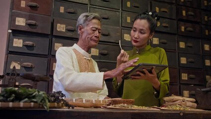 Wall Mural - Low angle view of young Asian daughter listening to aged father talking to her about traditional chinese herbal medicine near worktable at pharmacy