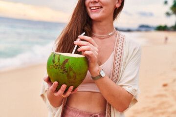 Boho girl traveler holds in hands a tropical green coconut with a straw on ocean shore