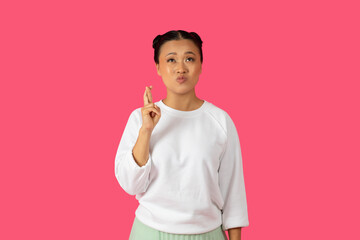 Asian woman with dark hair and a white shirt stands against a pink backdrop. She is crossing her fingers and blowing a kiss at the viewer.