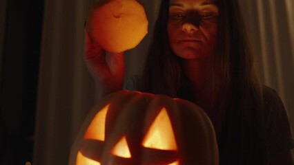 Wall Mural - In the Dark Room, the Woman Peeking Inside the Jack-o'-Lantern, Her Face Illuminated by Bright Warm Light from the Flame. She Closes It with the Lid. Slow Motion.