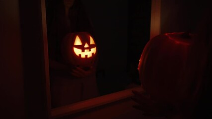 Wall Mural - The Woman Stands in Front of a Mirror in a Dark Room, Holding a Pumpkin with a Fire Inside the Jack-O'-Lantern, the Camera Moves Back.