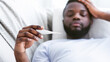 © Prostock-studio - Black man lays in bed, holding a thermometer in his hand. He is wearing a white t-shirt and looks uncomfortable, suggesting he might be sick.