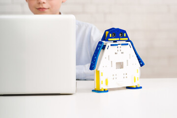 Aufkleber - Boy is working on a laptop computer with a toy robot sitting on the desk beside them. The robot is made of white plastic and has blue and yellow accents.