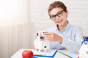 Poster - A young boy is sitting at a desk in his home, wearing glasses and a white shirt. He is smiling as he carefully assembles a white robot, focusing on the details of the construction.