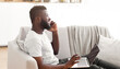 © Prostock-studio - Black guy entrepreneur working from home, sitting on couch with computer on his lap, have phone conversation, side view