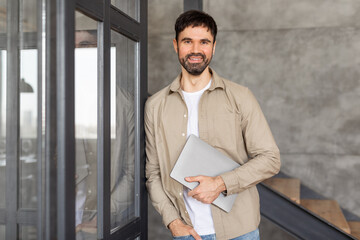 Poster - A man in a casual shirt stands in a modern office setting, holding a laptop computer close to his body. He is smiling at the camera while standing near a glass door with a view of the office interior.