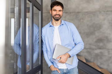 Poster - A man in a blue shirt and jeans stands in an office space, smiling and holding a laptop in his hands. He is looking directly at the camera, and his left hand is in his pocket.