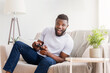 © Prostock-studio - A young black man sits on a couch in his living room, excitedly playing video games using a black controller. He is wearing a white t-shirt and jeans, and there is a plant on a side table behind him.