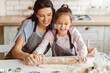 © Prostock-studio - A mother and daughter are baking together in a kitchen. The young girl is rolling out dough with a rolling pin while her mother helps guide her hand. They are both wearing aprons and smiling.