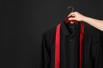 Woman holding hanger with shirt and necktie on black background, space for text