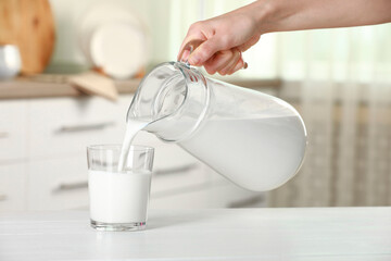 Wall Mural - Woman pouring fresh milk from jug into glass at white wooden table indoors, closeup