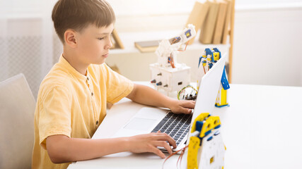 Aufkleber - A young boy is learning about robotics by using a laptop computer. The boy is sitting at a table and is looking at the screen of the laptop. There is a robotic arm behind the laptop.