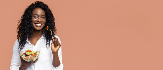 African American woman with curly black hair smiles as she eats a fresh salad with a fork. She is wearing a white shirt and stands against a peach-colored background.